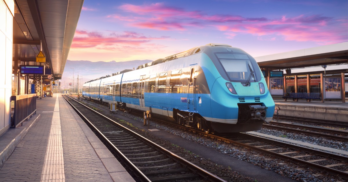 A blue high-speed train on a railway at an empty public transport station. The sun sets behind the train.