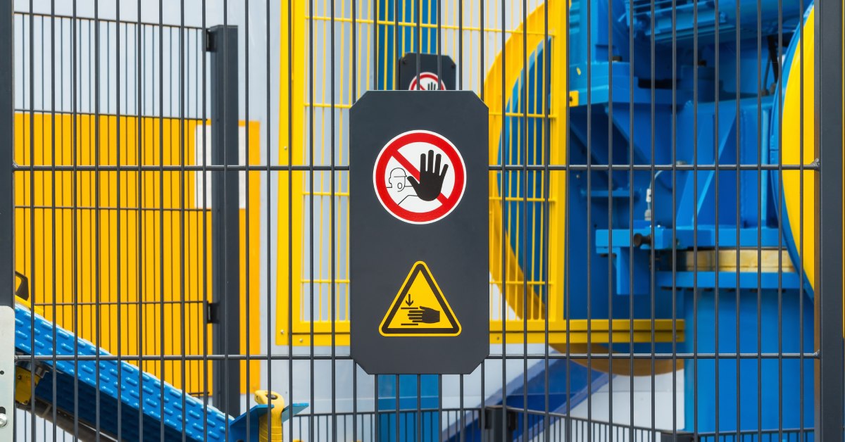 A machine guarding fence separates equipment from workers, with a safety sign showing symbols to not touch it.