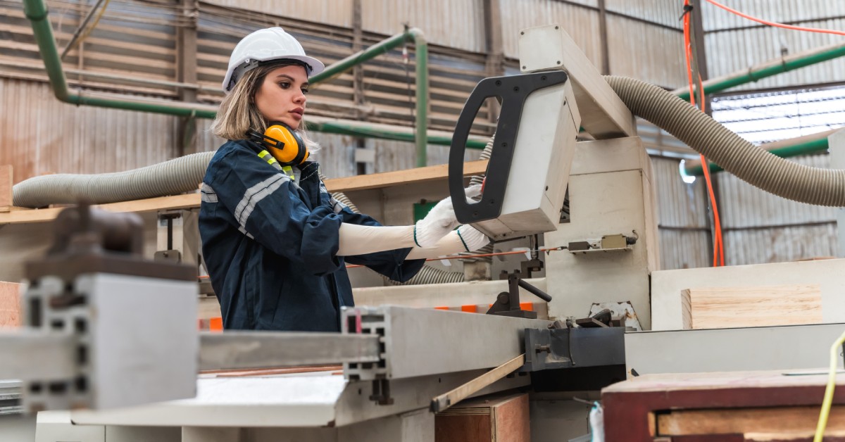 An engineer wearing a white hard hat and navy jacket operates a manufacturing machine in a warehouse.