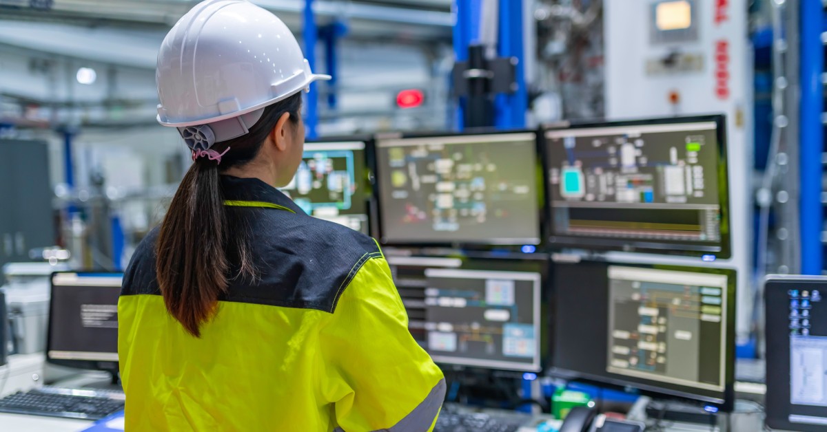 An engineer in a yellow jacket and white hard hat stands in front of a desk with multiple monitors and keyboards.