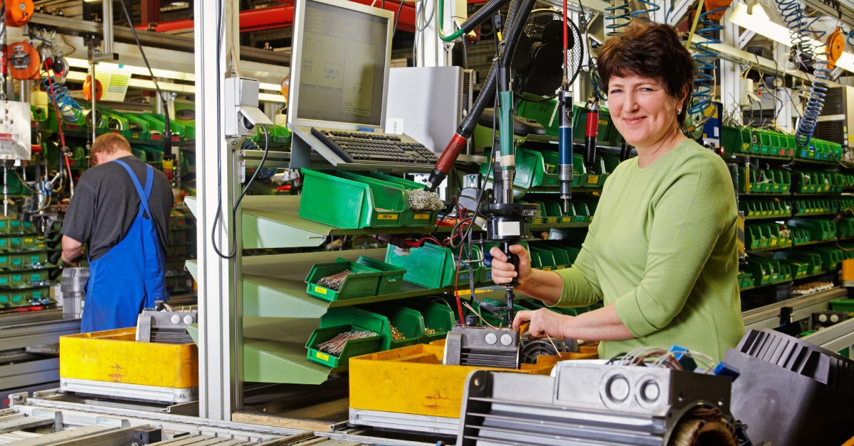 A woman stands at an industrial workbench while assembling a product near multiple containers filled with parts.