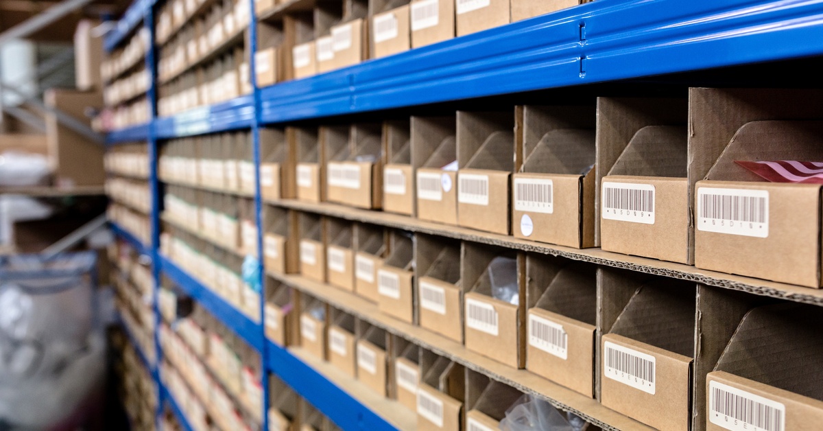 A long shelf in a warehouse with rows of small cardboard boxes containing items the factory needs to store.