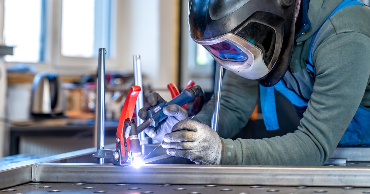 A metalworker is leaning over a workbench, welding pieces of metal together while wearing protective gear.