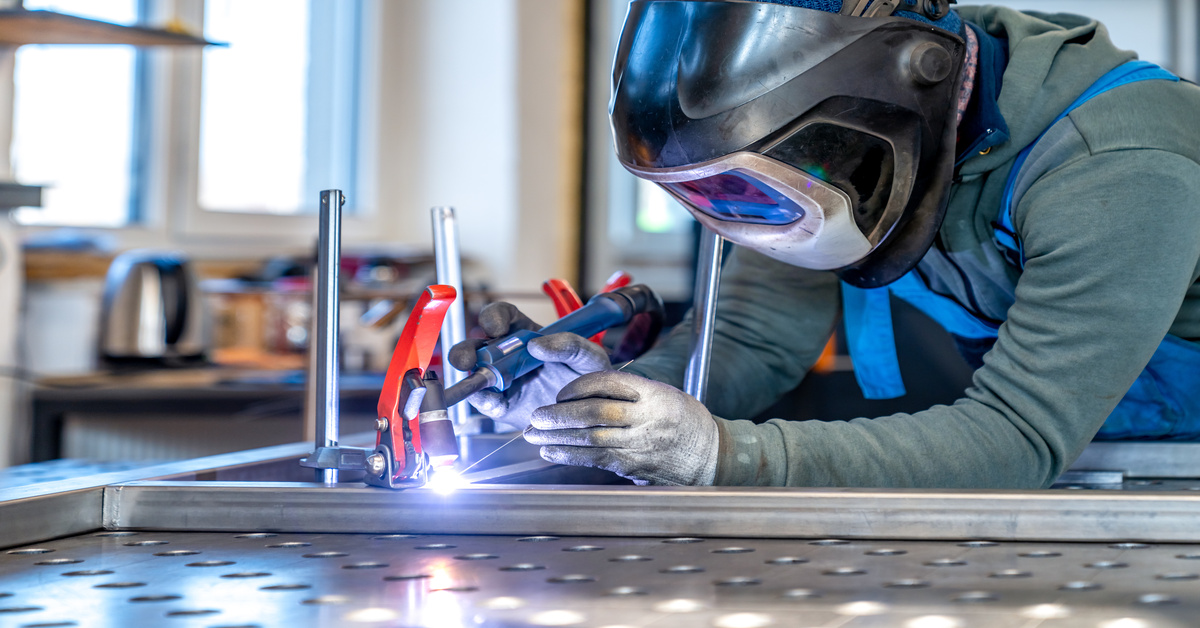 A metalworker leaning over a frame in a workshop and attaching two pieces of metal. He is wearing protective gear.