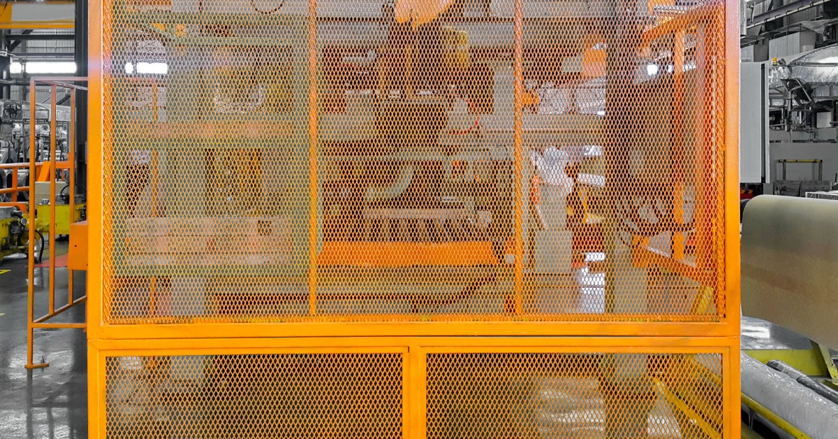A large safety guard fence surrounds a complex piece of machinery in an industrial factory that has other machines.