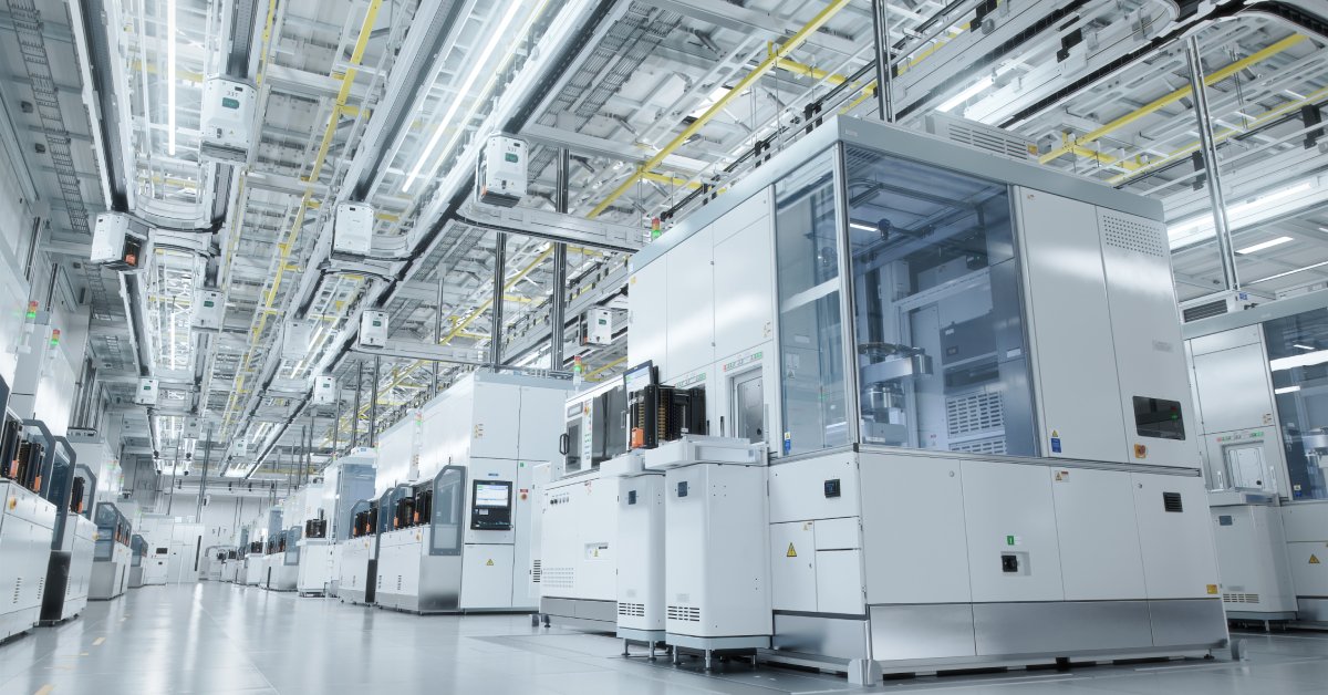 A brightly lit cleanroom in a facility. Inside the cleanroom, large machines are lined up in a row, working.